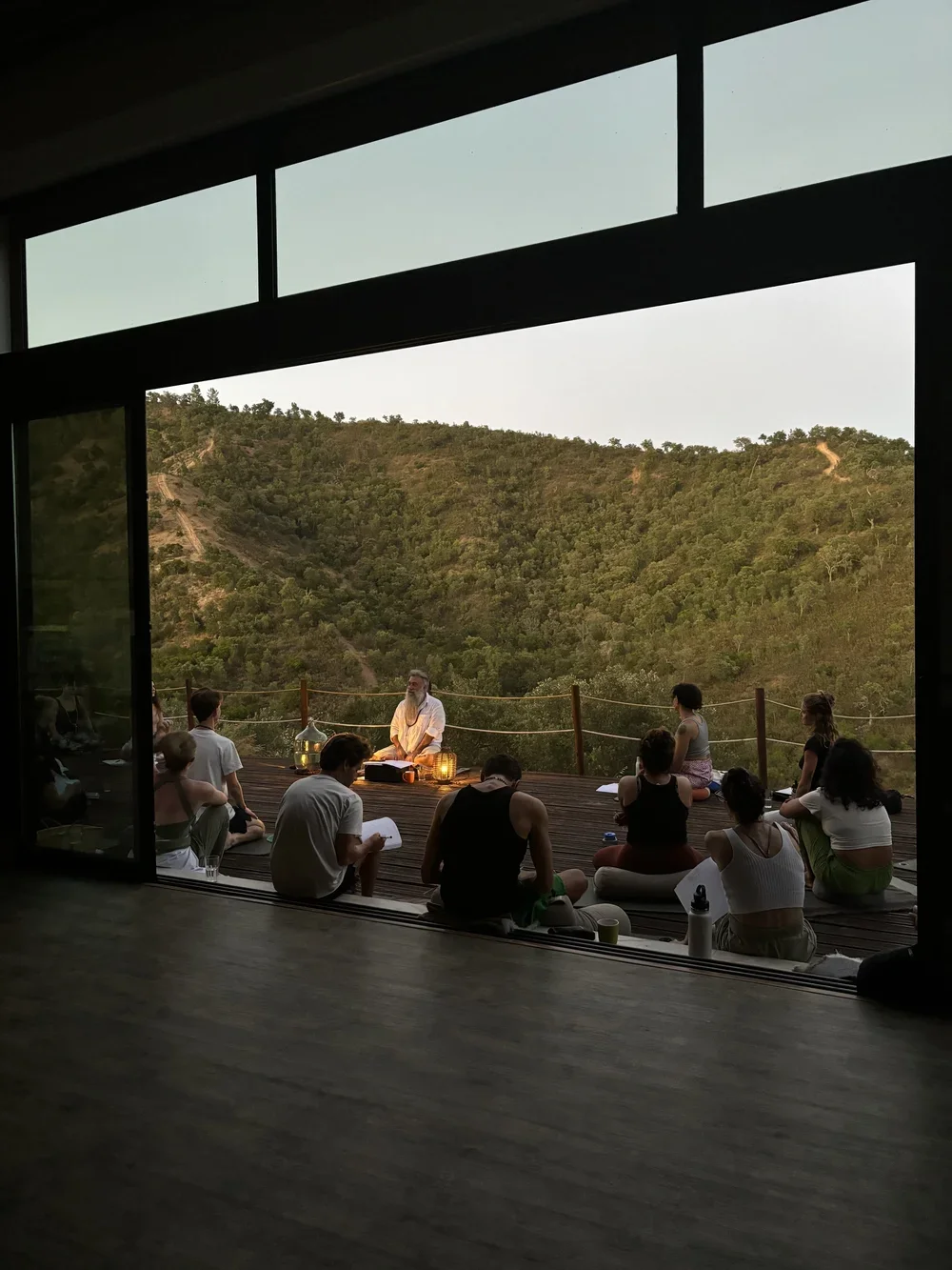 Grupo de personas sentadas en un deck de madera en un entorno natural, participando en una sesión de meditación, con un instructor en el centro, rodeados de naturaleza y montañas, en retiro