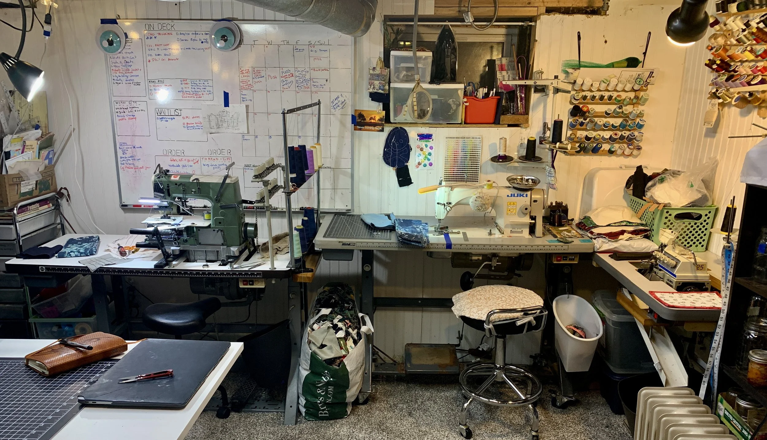 a behind-the-scenes photo of a cozy (cramped) basement studio workspace with several industrial sewing  machines and racks of thread
