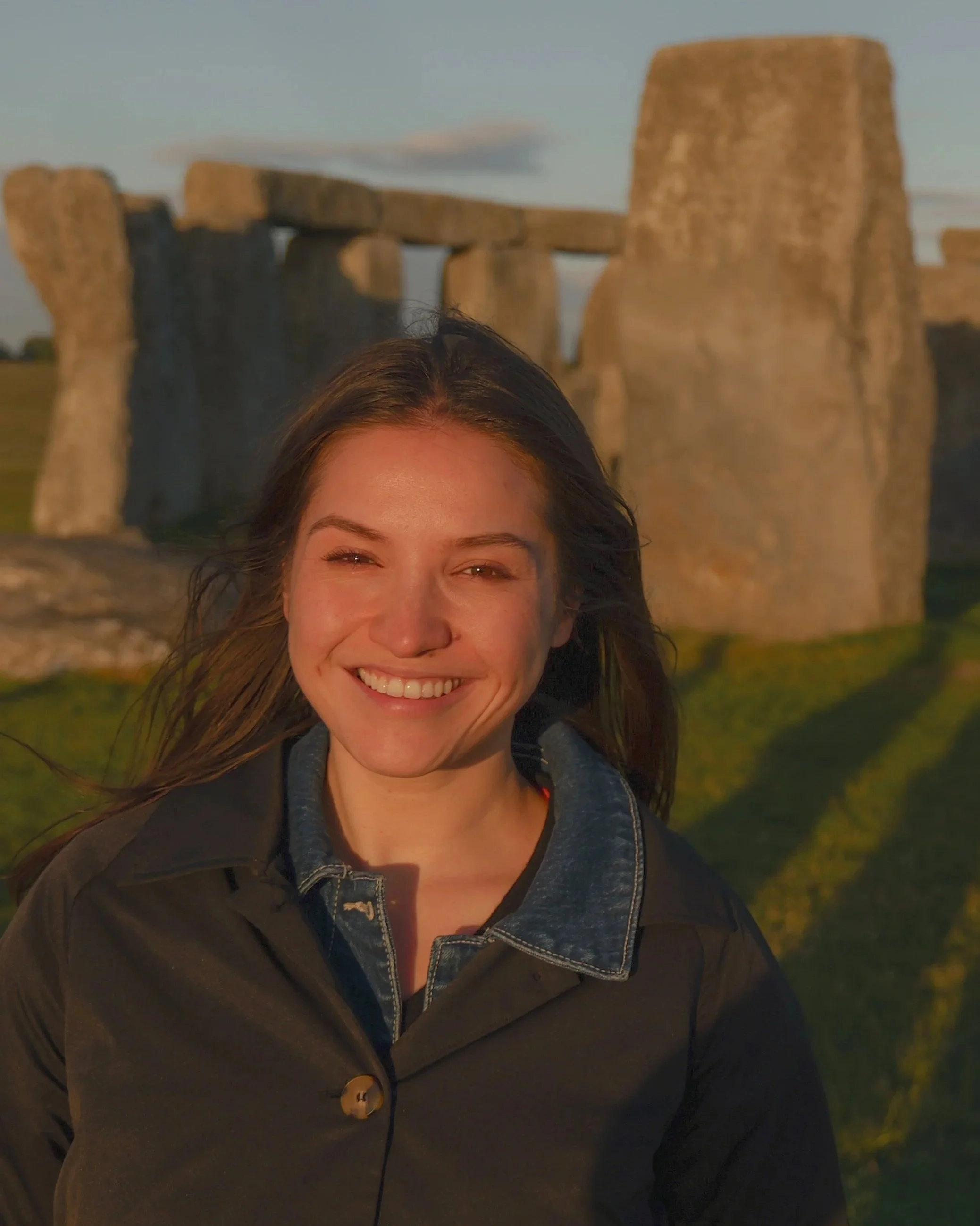 Close-up of a smiling woman with dark hair outdoors near Stonehenge during sunset