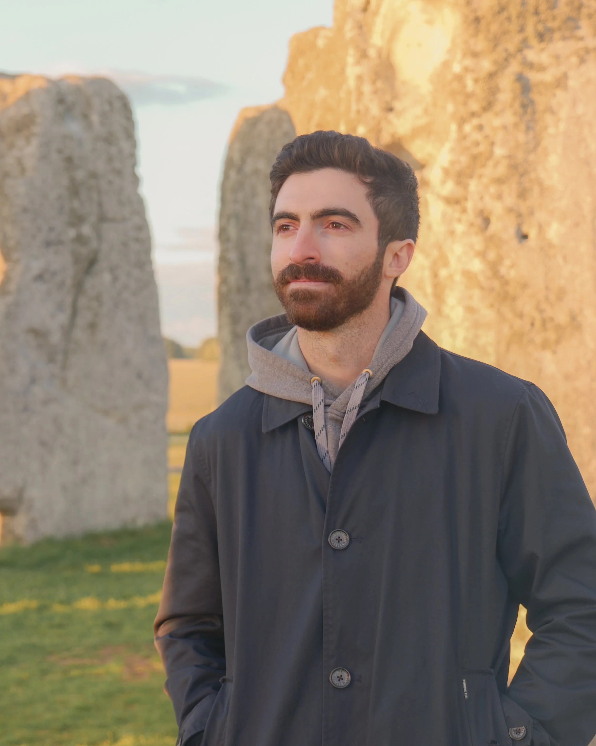 A man with dark hair and a beard stands outdoors during sunset near Stonehenge, wearing a dark jacket and a gray hoodie.