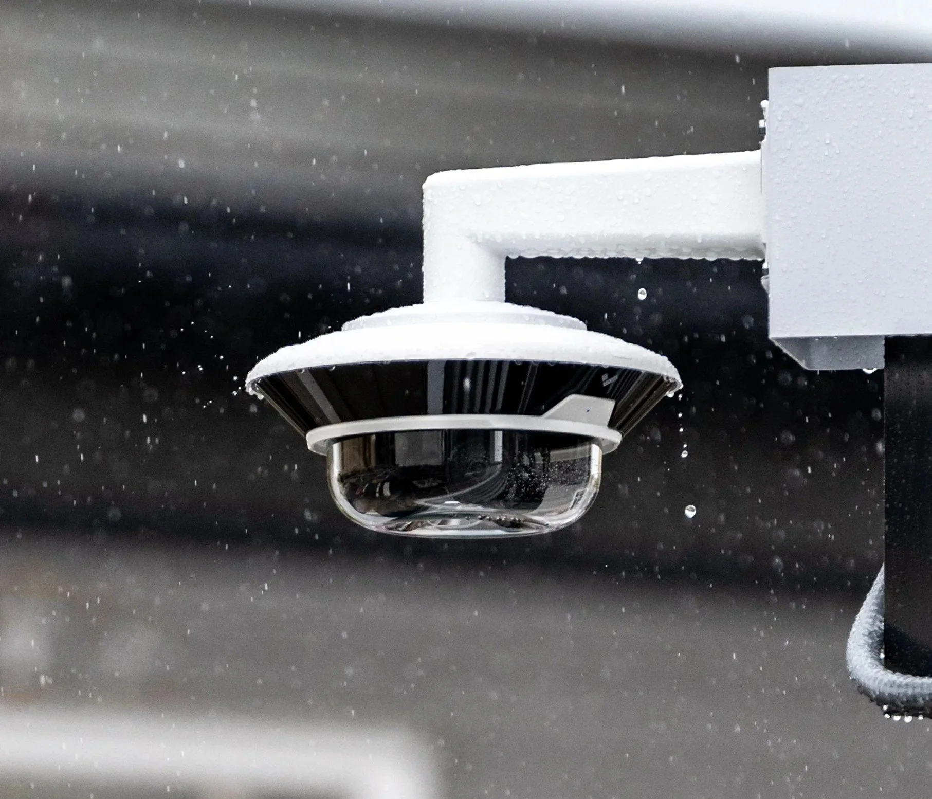 Close-up of a security camera mounted on a black surface with water droplets, with rain falling in the background.