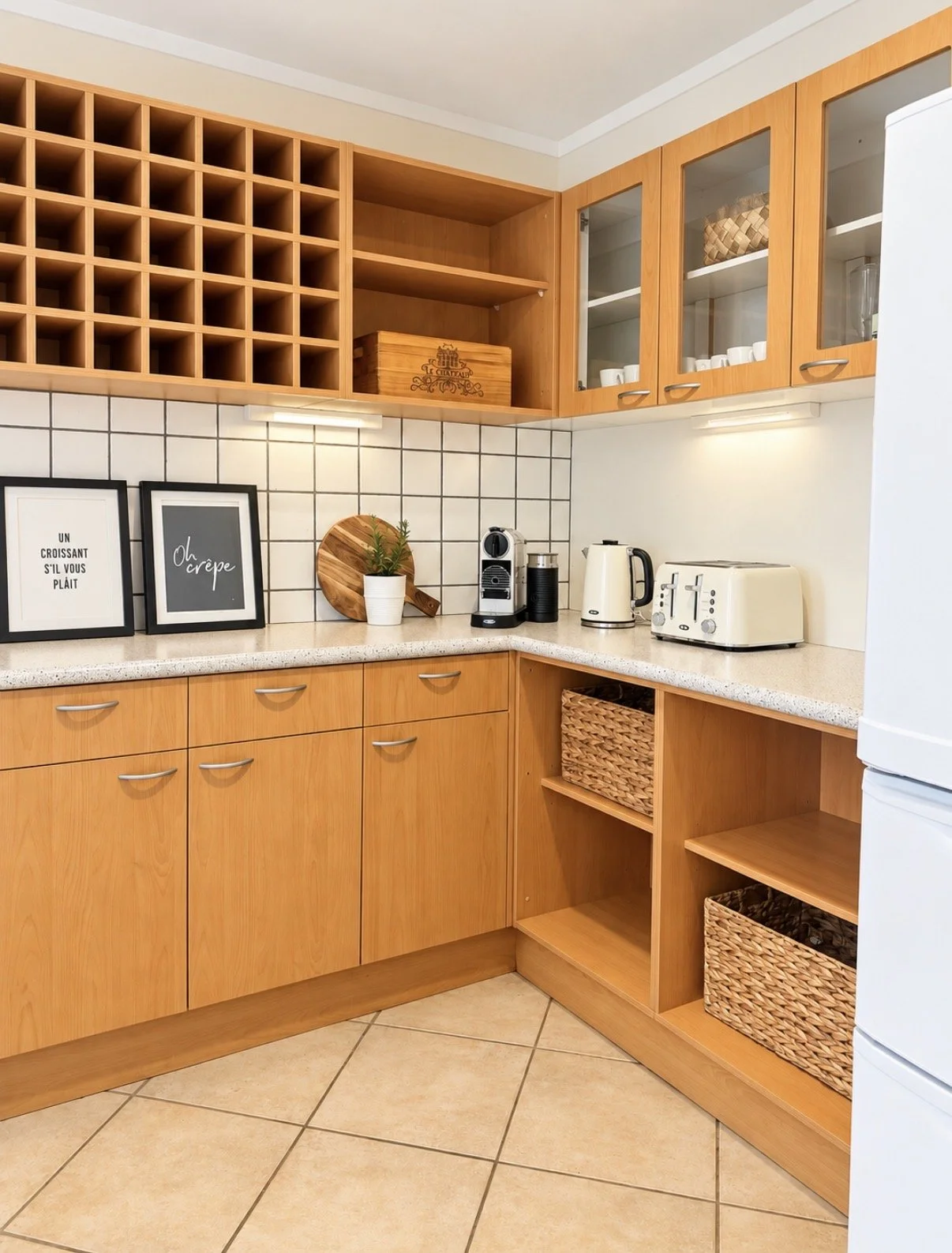 A kitchen with wooden cabinets, open shelves, black and white framed quotes, a small potted plant, and kitchen appliances including a coffee machine, kettle, and toaster. There are woven baskets in open cubbies and glassware in the upper cabinets.