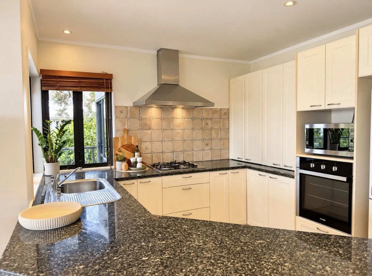Modern kitchen with beige cabinets, black granite countertops, a tiled backsplash, a stainless steel range hood, a stove, a microwave, a potted plant, and a window with brown blinds.