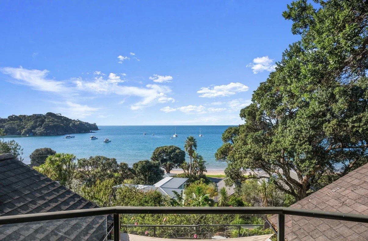View of a beach and ocean from a balcony with rooftops, trees, and boats in the water under a partly cloudy sky.
