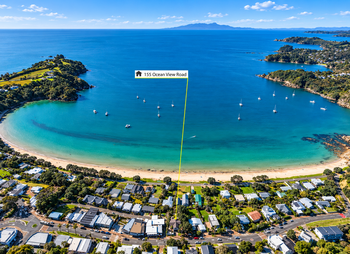 Aerial view of a coastal neighborhood with houses, a sandy beach, and a bay with sailboats. The water is clear and blue, with a small green island in the bay. The horizon features distant mountains under a partly cloudy sky.