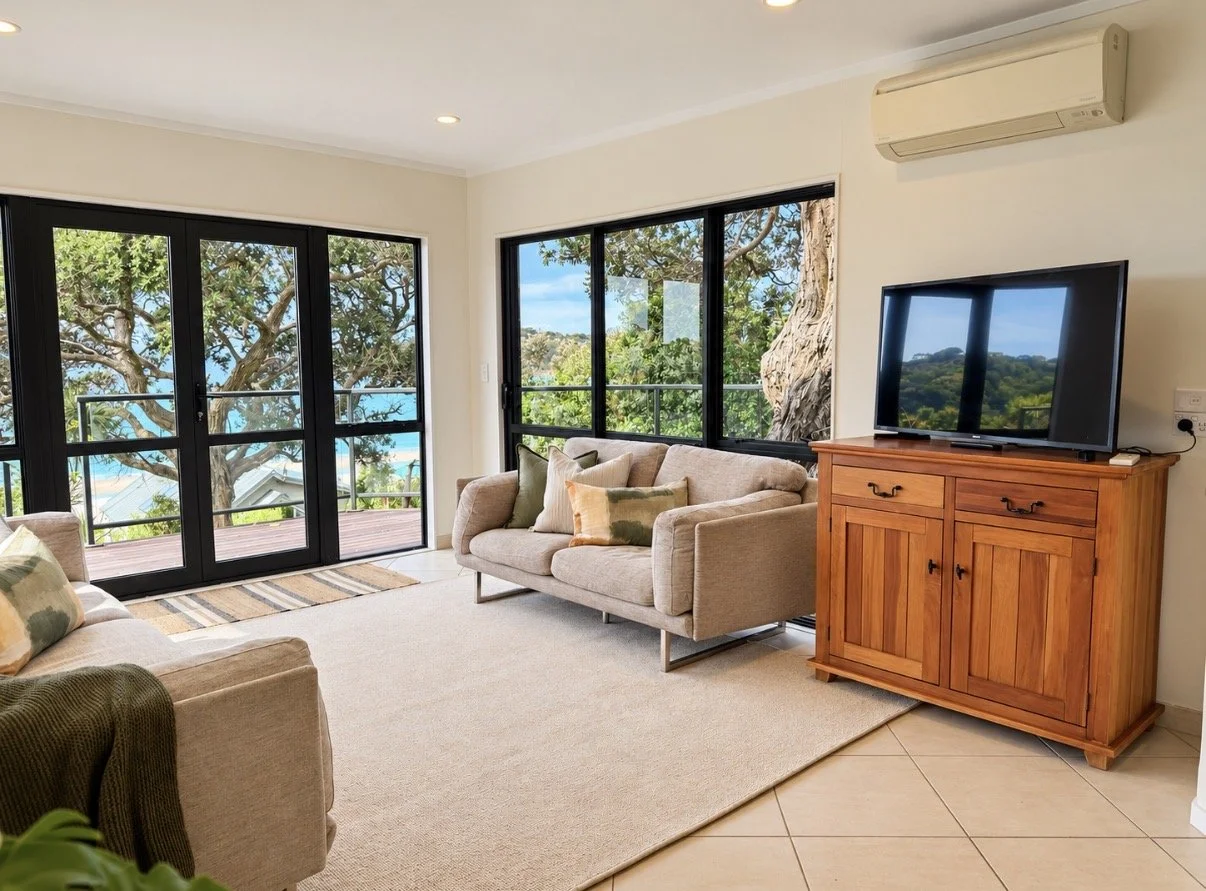 Living room with large windows, beige couches with pillows, wooden cabinet with television, air conditioning unit, glass sliding doors leading to a balcony, view of trees and blue sky outside.