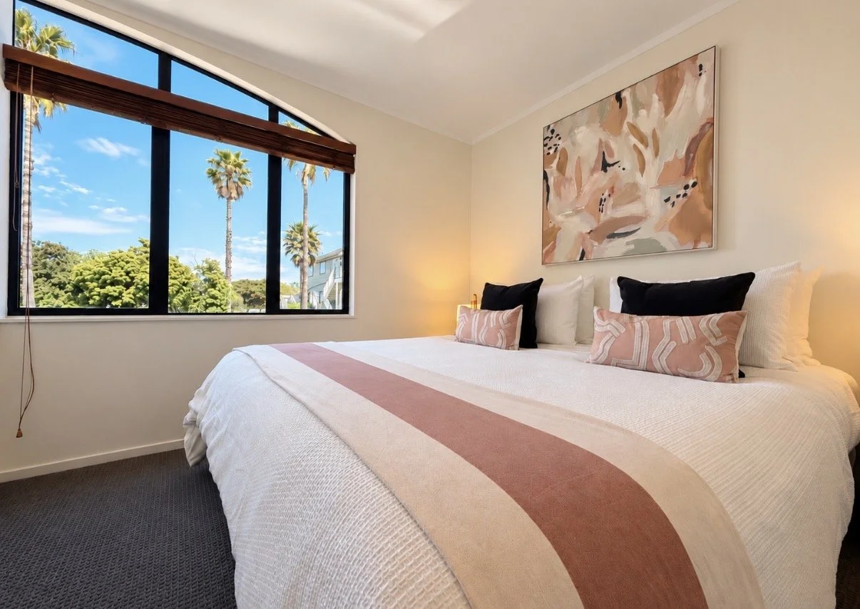 A bedroom with a large bed covered in white bedding with a pink and beige striped throw. There are four pillows, two black and two pink patterned. An abstract floral painting hangs above the bed. A large window reveals tall palm trees and a blue sky 
