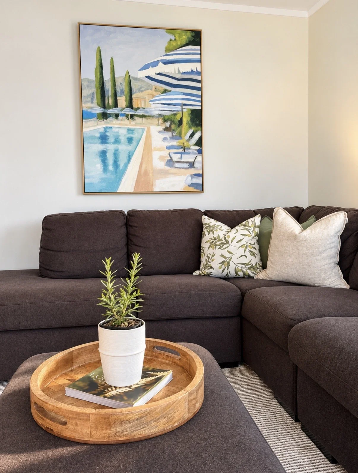 Living room with a dark brown sectional sofa decorated with floral and plain pillows, a wooden tray table with a potted plant and a magazine, a large landscape painting of a pool with umbrellas and tall trees on the wall, and a striped rug.