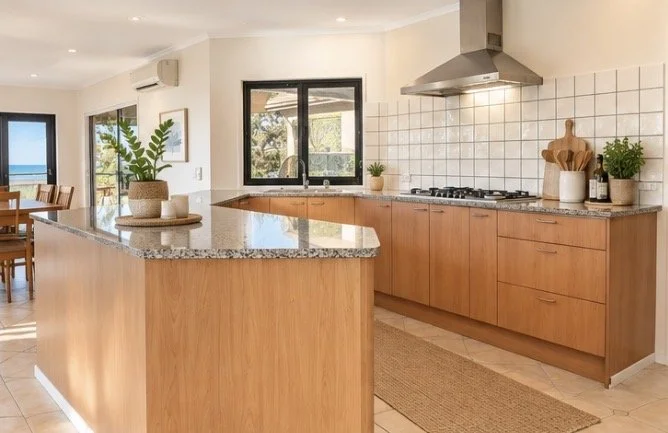 Modern kitchen with light wooden cabinetry, granite countertop island, white tiled backsplash, stainless steel stove hood, and potted plants near window.