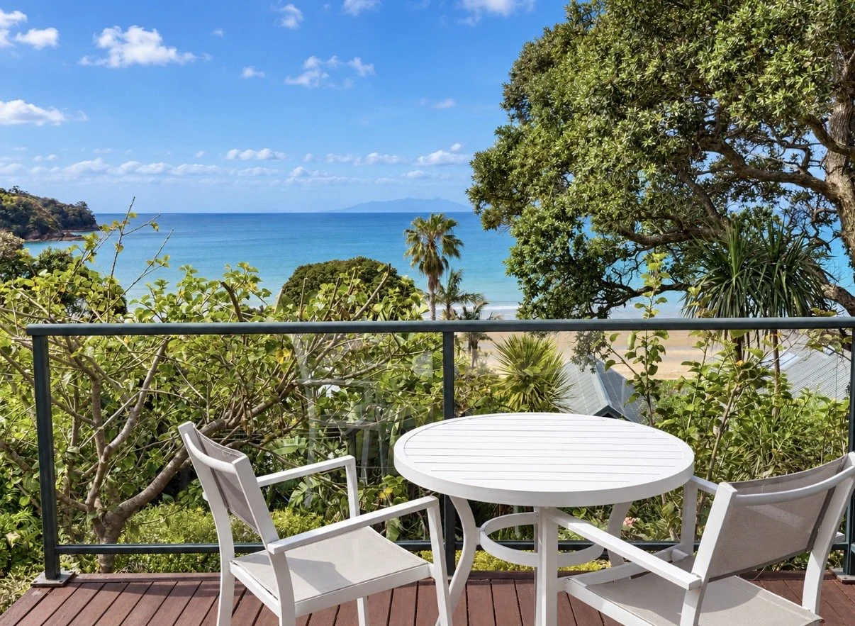 Balcony with a white table and chairs overlooking a lush garden, sandy beach, and ocean with blue sky and clouds.