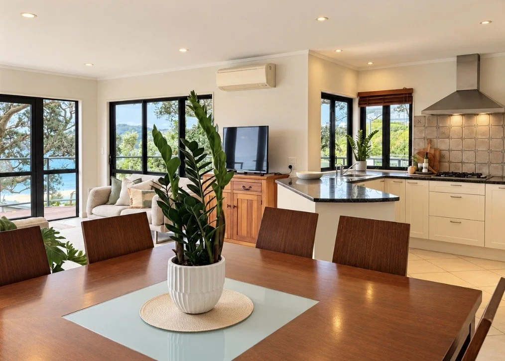 Open-concept living and kitchen area with large windows, a wooden dining table with a potted plant, modern white kitchen cabinets, black countertops, and a view of outdoors with trees and water