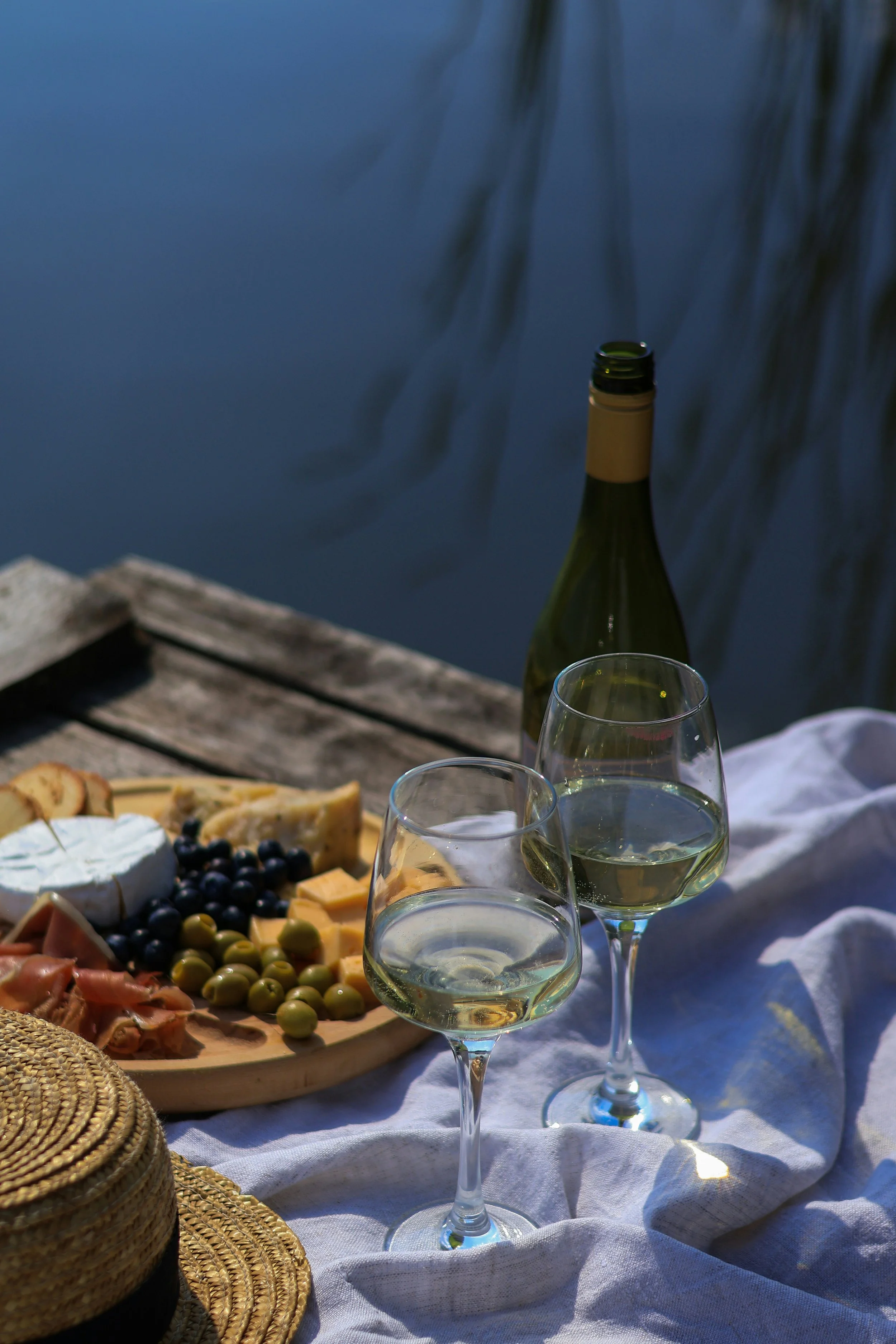 A picnic table with a cheese platter, grapes, and two glasses of white wine, set outdoors near a body of water with some greenery in the background.