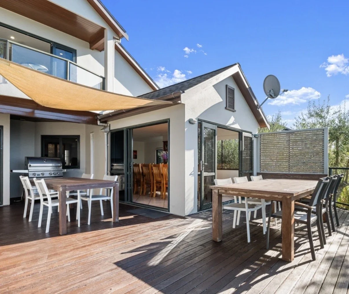 Outdoor patio with wooden dining tables and chairs, a gas grill, sliding glass doors, and a blue sky with some clouds.