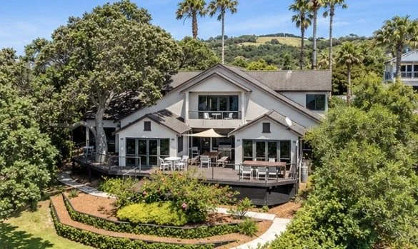 A house with multiple large windows and a spacious deck, surrounded by lush greenery and tall palm trees, with a garden and a pathway in front.