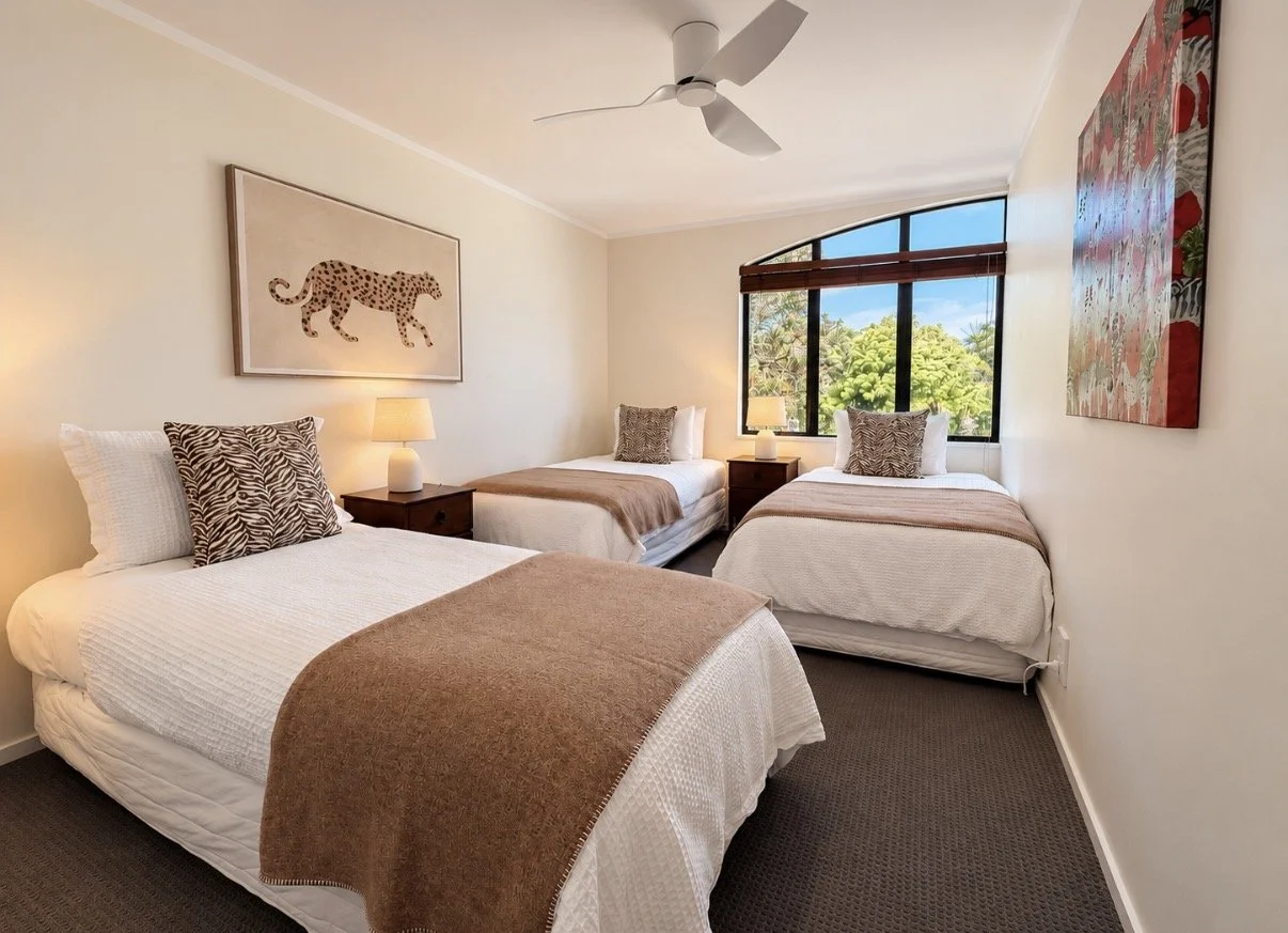 A bedroom with three beds, each with white bedding, patterned pillows, and brown throw blankets. There are bedside tables with lamps, a ceiling fan, a large window with a view of greenery outside, and wall art including a cheetah picture and a colorf