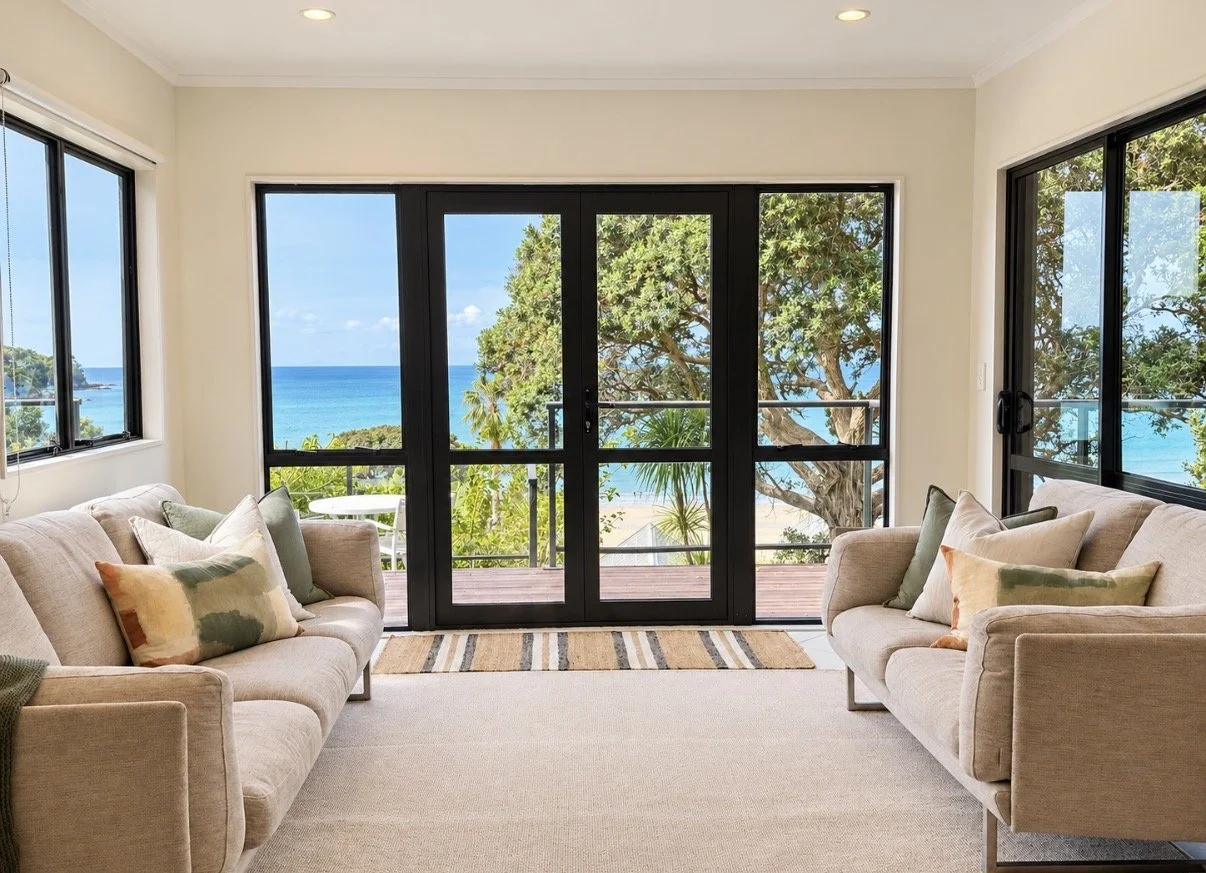 Living room with beige sofas, colorful pillows, and large sliding glass doors leading to a balcony with a view of the ocean and trees.
