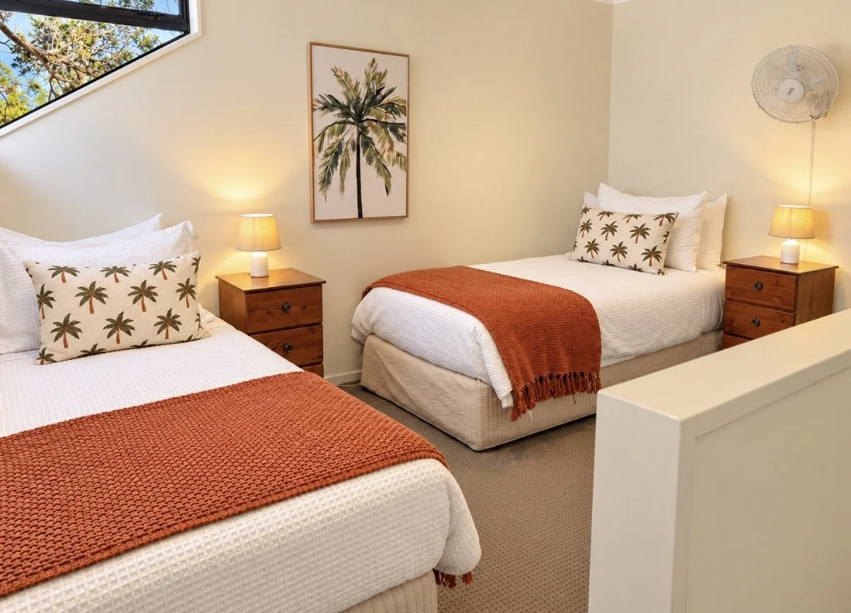 A bedroom with two beds, each decorated with patterned pillows featuring palm trees, and orange throws at the foot of each bed. There are wooden nightstands with lamps between and beside the beds. A framed picture of a palm tree hangs on the wall, an