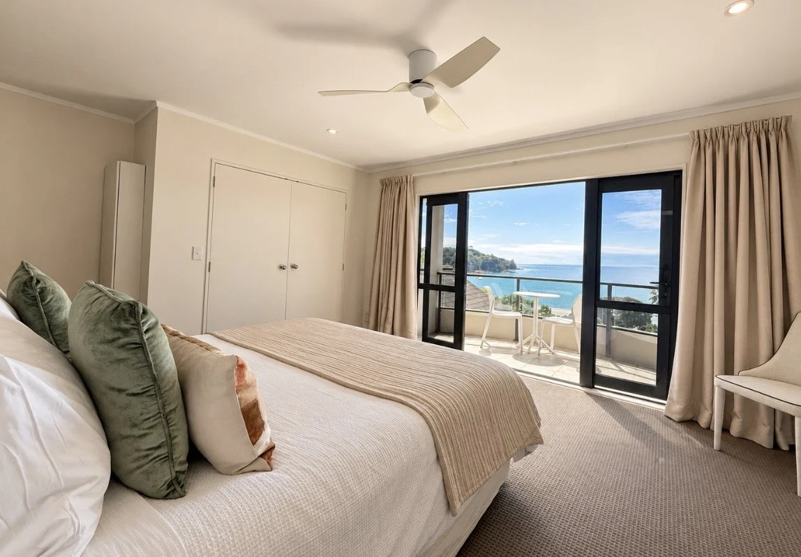 A bedroom with a large bed, green and beige pillows, and a beige blanket, facing a glass sliding door with a view of a balcony overlooking the sea and distant land.