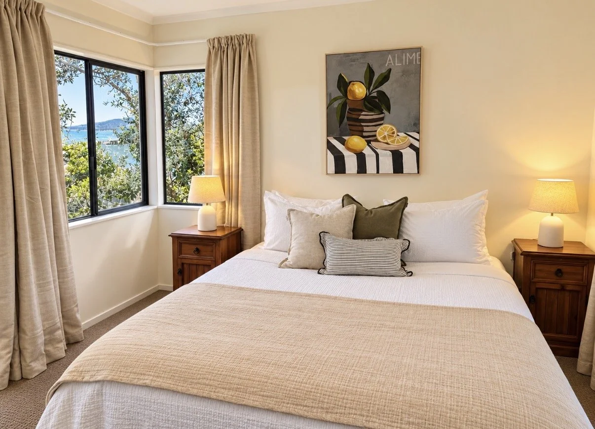A cozy bedroom with a bed, multiple pillows, bedside tables with lamps, large windows with curtains, and a colorful fruit-themed artwork hanging above the bed.