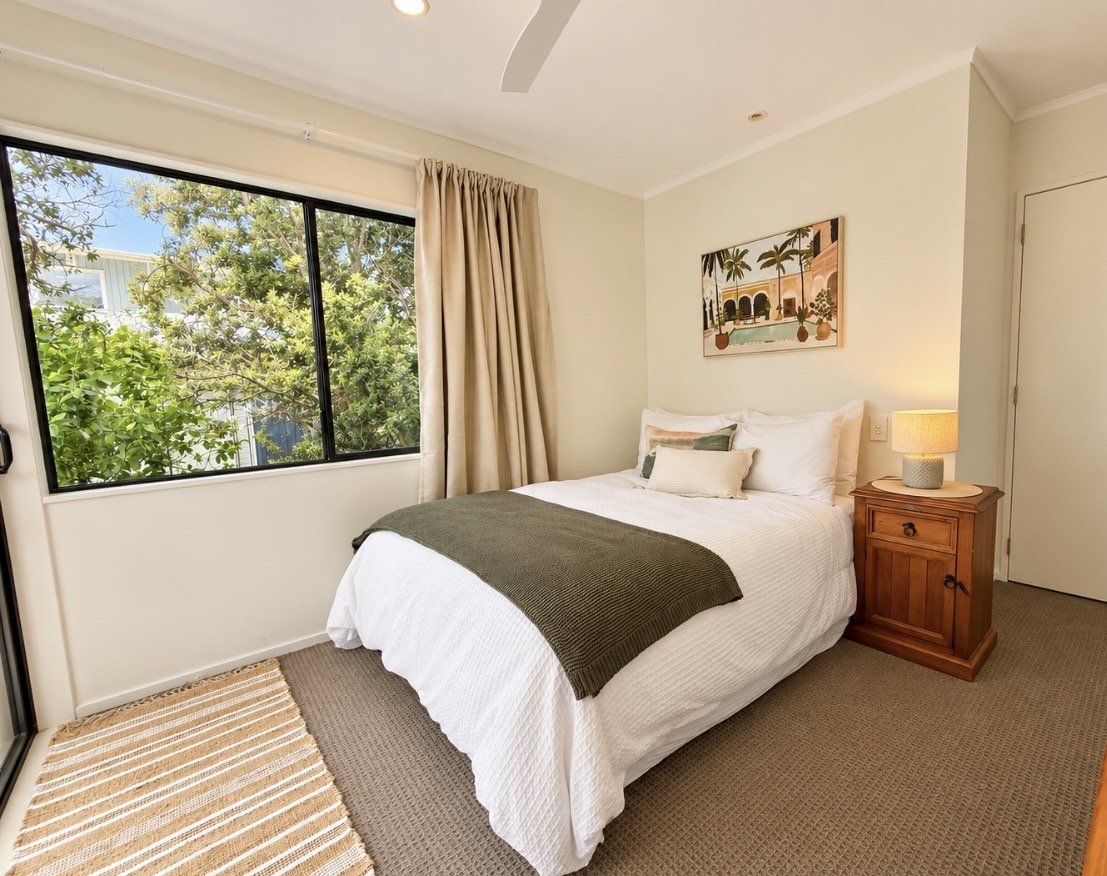 A bedroom with a large window showing trees outside. The bed has white bedding, a green throw blanket, and multiple pillows. A wooden nightstand with a lamp is next to the bed. There's a framed picture of a tropical scene on the wall above the bed.