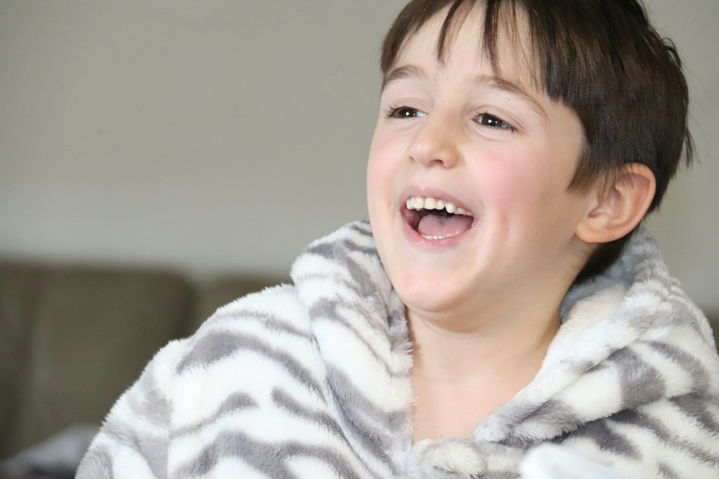A young boy with brown hair, smiling and showing his teeth, wearing a fuzzy striped hoodie.