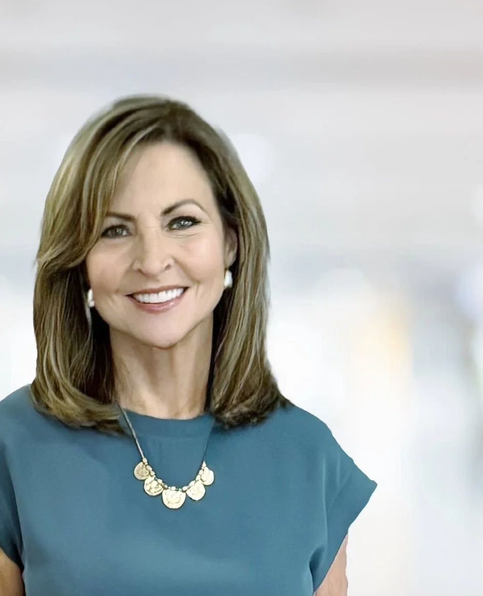 A smiling woman with shoulder-length brown hair wearing a teal top, pearl earrings, and a gold necklace with circular pendant charms.
