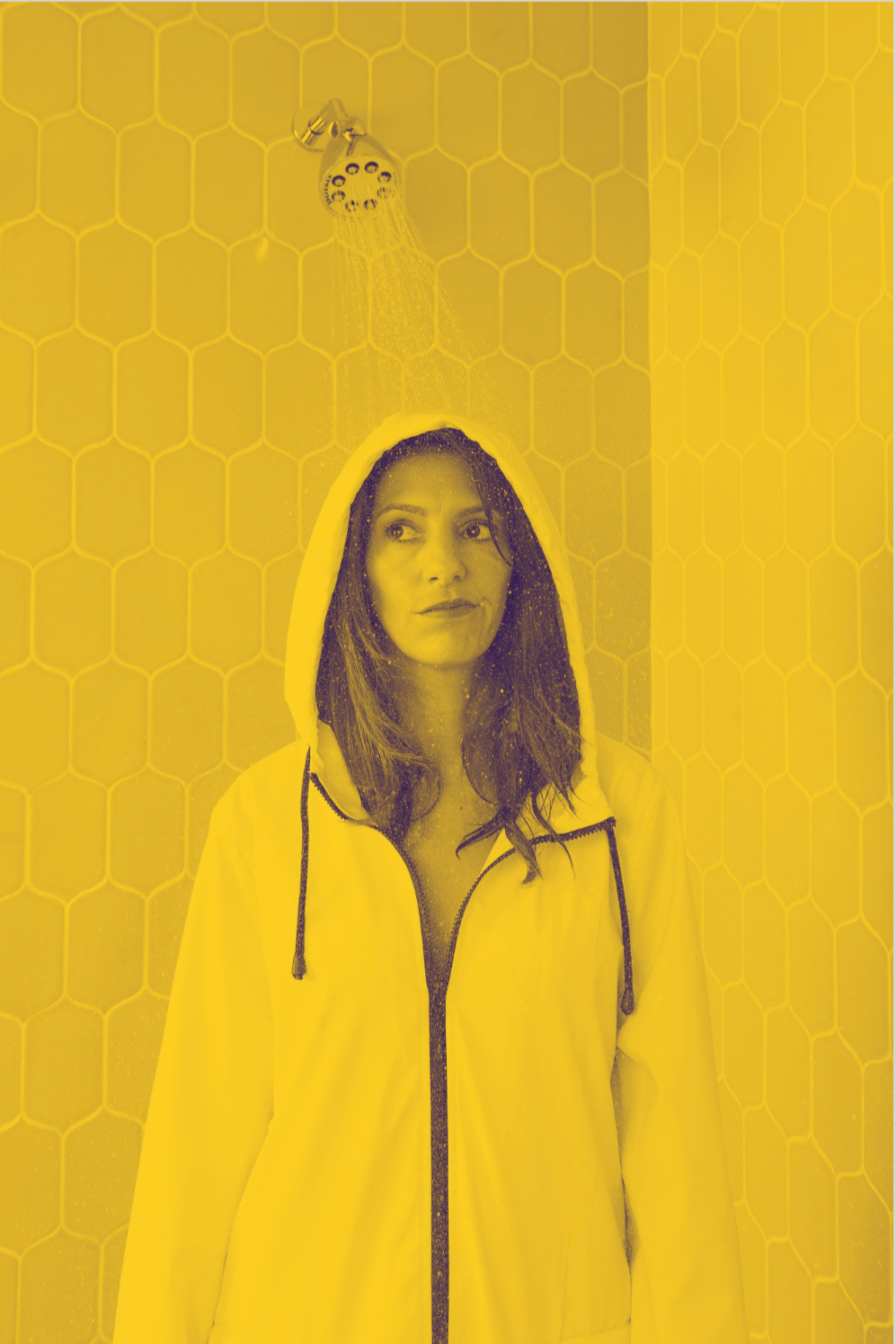Clara Bijl with long brown hair wearing a yellow raincoat is standing under a shower in a room with yellow honeycomb-patterned tiles.