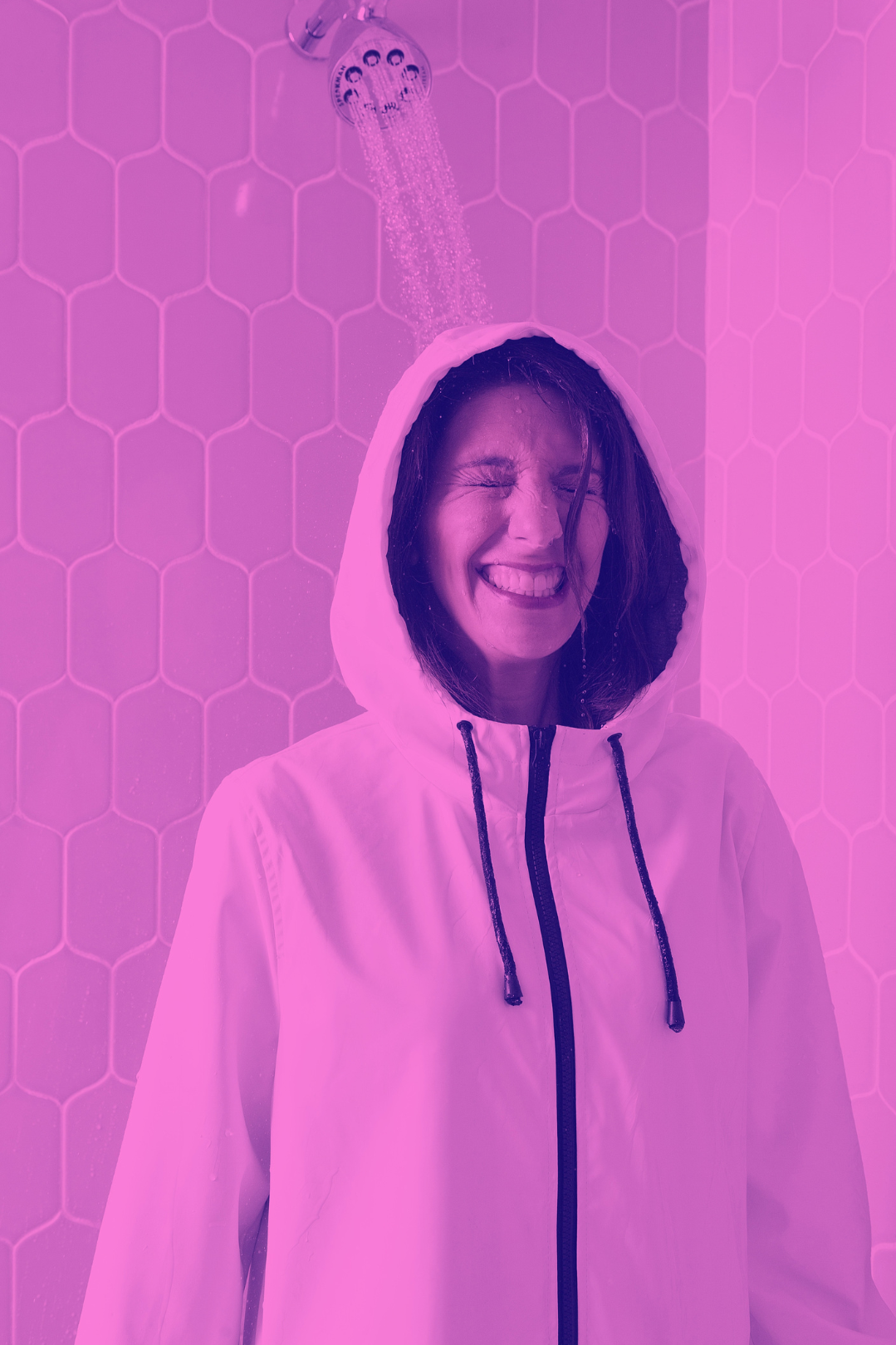 Clara Bijl wet hair smiling and squinting under a showerhead, wearing a white hooded jacket, in a bathroom with white hexagonal tiles on the wall, bathed in pinkish-purple light.