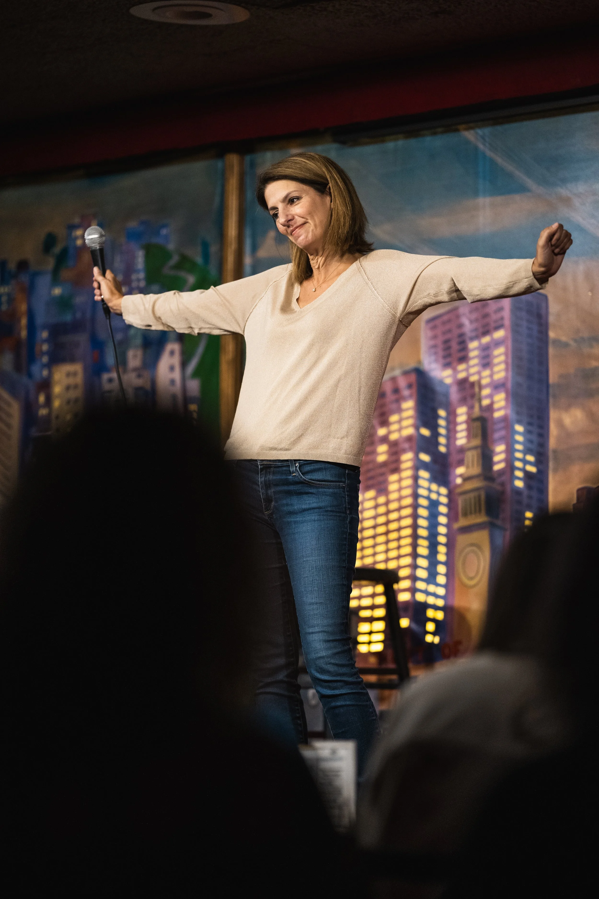 A woman with shoulder-length brown hair standing on a stage, holding a microphone in her right hand, wearing a beige long-sleeve shirt and jeans, with a cityscape backdrop painted on the wall behind her.