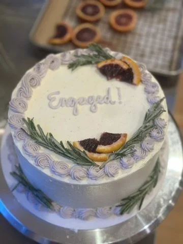 white and purple cake that reads "engaged" with sugar-covered blood orange and rosemary sprigs