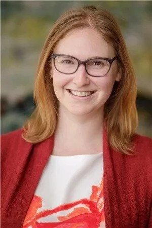 A woman with red hair, glasses, and a red blazer smiling outdoors.