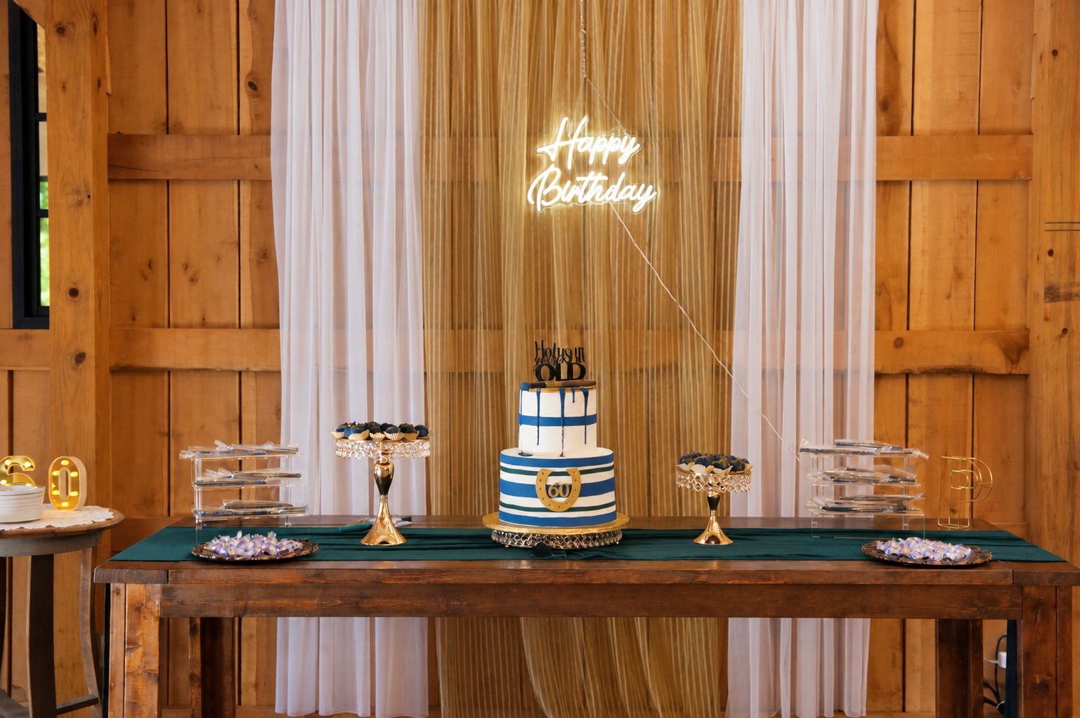 Styled 60th birthday cake table with gold dessert stands and navy, emerald and gold accents.