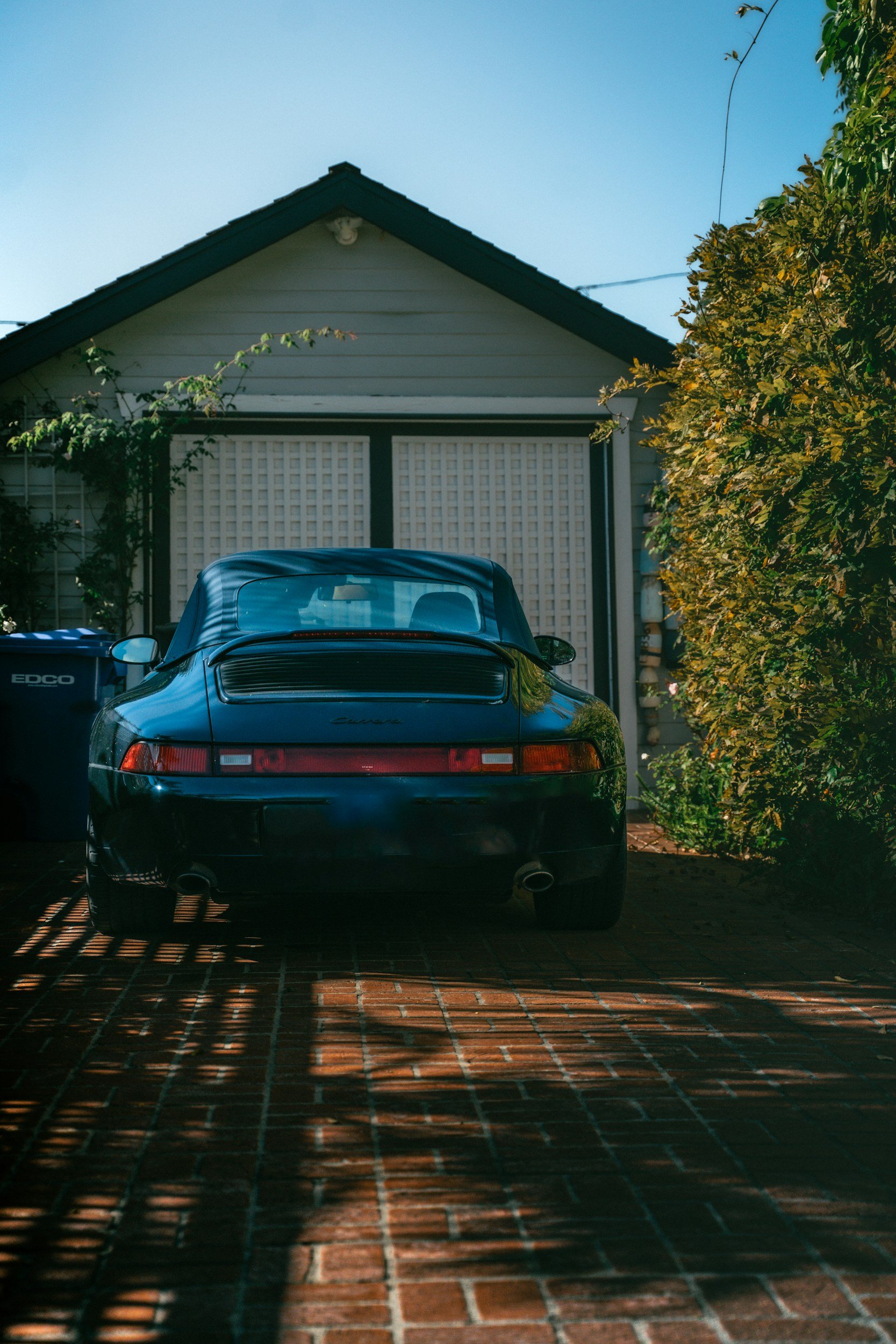 A black Porsche Carrera parked on a brick driveway in front of a house with a gabled roof, surrounded by green shrubs and trees, under a clear blue sky.
