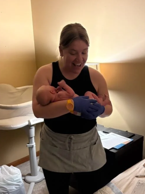 A woman holding a newborn baby in a hospital room, smiling and looking at the baby.