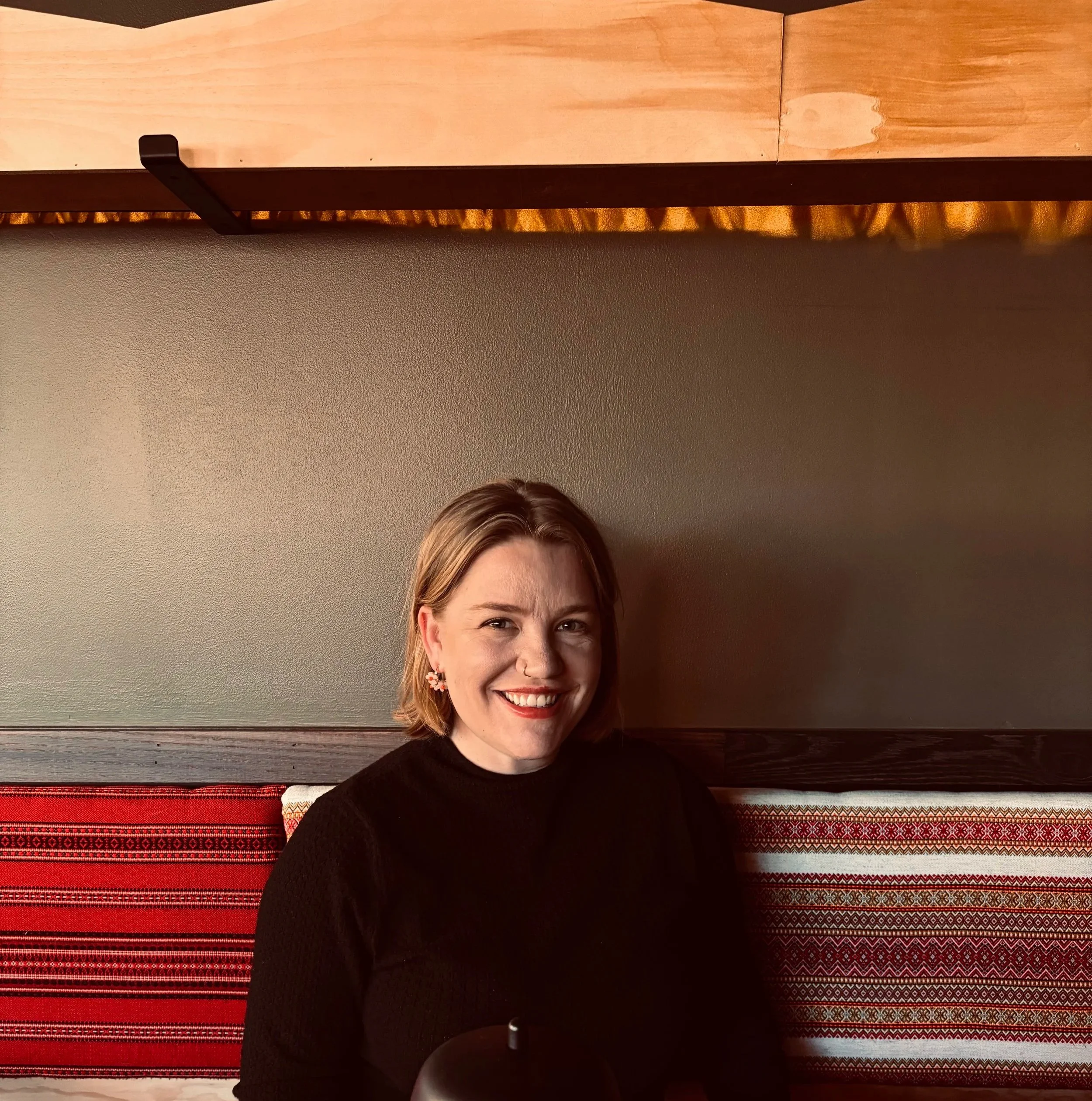 A smiling woman with light brown hair, wearing earrings, sitting in front of a beige and red woven blanket and a brown wooden wall.