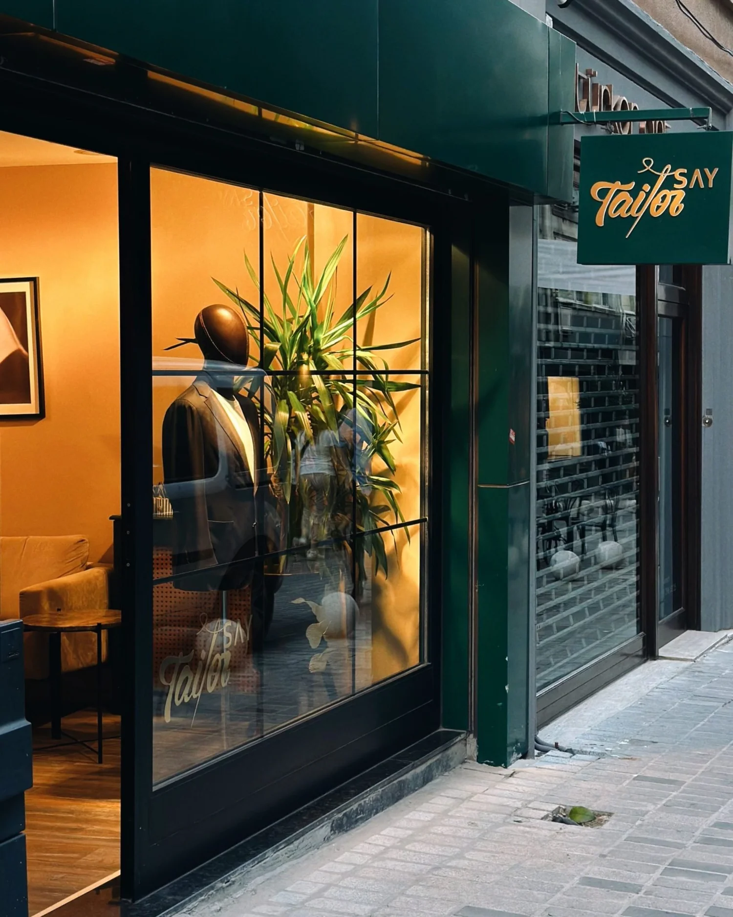Warmly lit tailor shop window with a mannequin and a plant at dusk