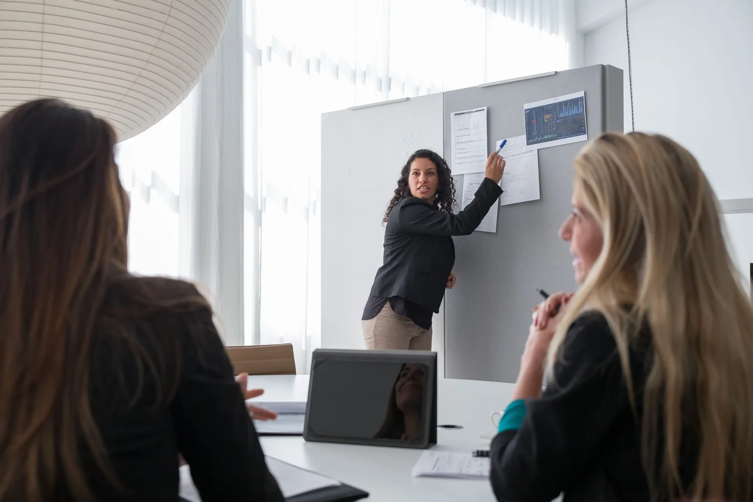 A woman presenting to a small group of seated colleagues in front of a gray presentation board