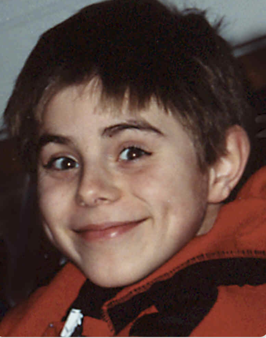 A young boy with short brown hair and blue eyes smiling at the camera, wearing an orange and black jacket.