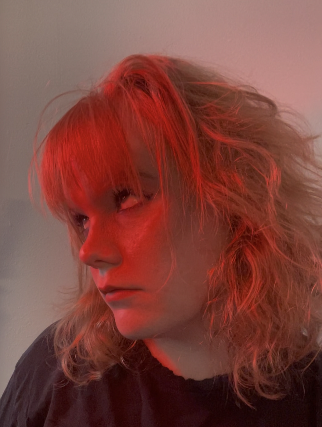 Close-up of a young woman with wavy blonde hair, illuminated by red lighting, wearing a dark top, against a neutral background.