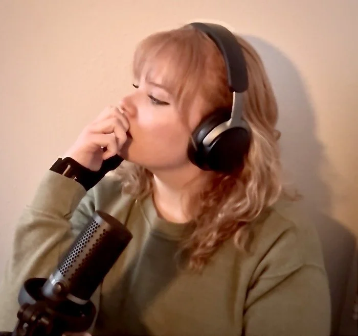 A young woman with red hair wearing headphones, sitting at a microphone in a recording studio.