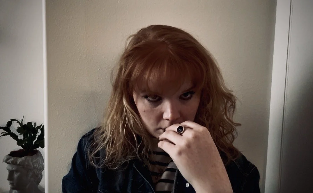 A woman with red hair and freckles looking at the camera, wearing a black ring and a striped shirt, sitting indoors in front of a beige wall.