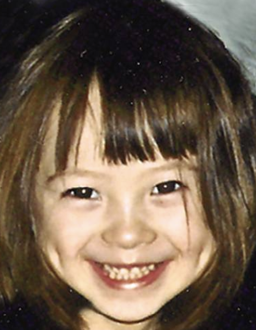 Close-up photo of a young girl with brown hair and bangs smiling broadly showing her teeth.