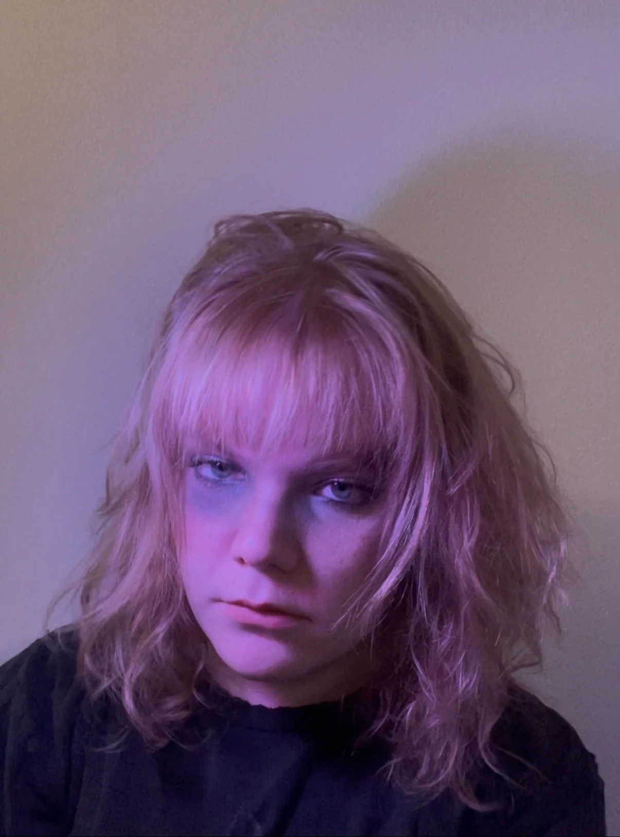Close-up of a young woman with pastel pink hair, blue eyes, and a black shirt, looking at the camera with a neutral expression.