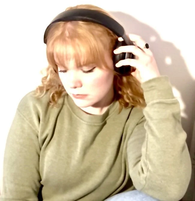 Young woman with red hair wearing a green sweatshirt and headphones, looking down with a calm expression.