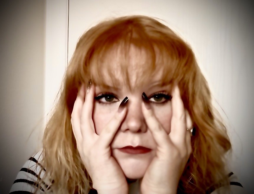 A woman with red hair and makeup holding her face with both hands.