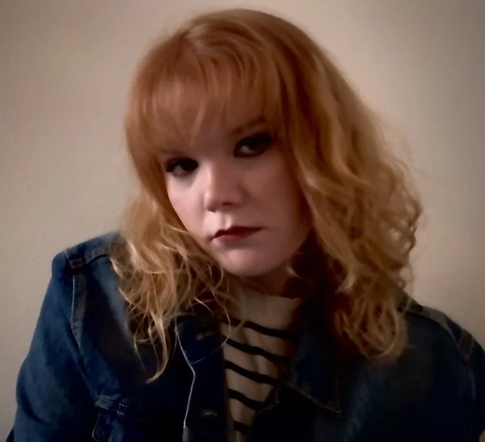 A woman with shoulder-length, curly, red hair and dark makeup, wearing a striped shirt under a denim jacket.