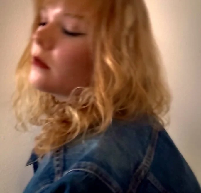 A young woman with curly blonde hair wearing a denim jacket, looking down with a soft expression.