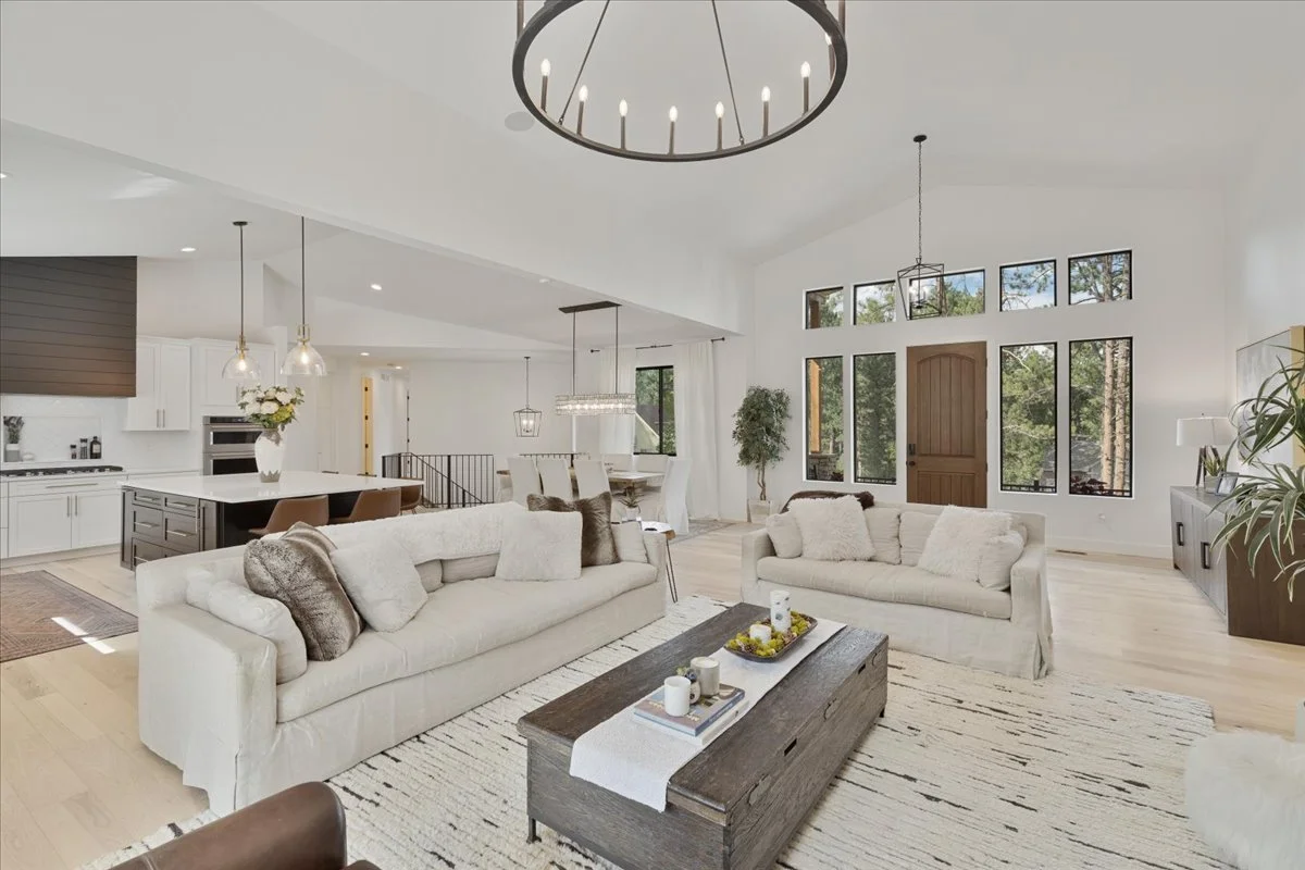 Bright open-concept living room and kitchen featuring vaulted ceilings, white sofas, a large kitchen island, black-framed windows, and light hardwood flooring