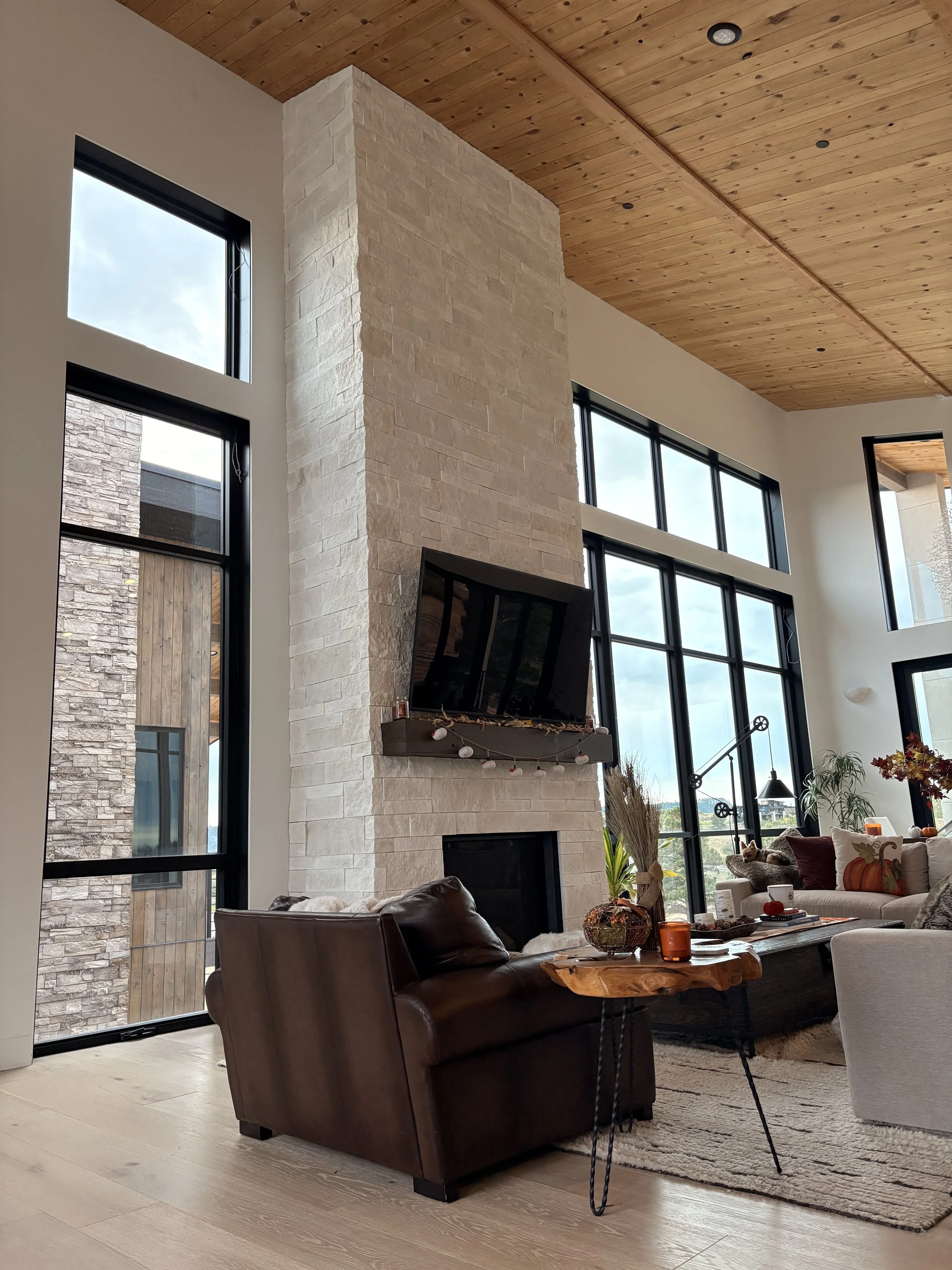 Floor-to-ceiling stone fireplace in a modern living room featuring a vaulted wood ceiling, tall black-framed windows, and natural light.