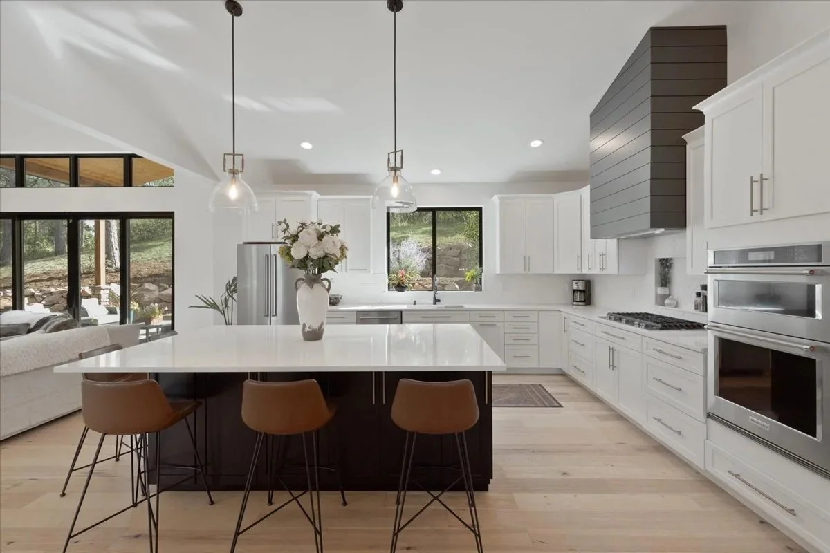 Open-concept kitchen featuring white shaker cabinets, a dark wood island with seating, stainless steel appliances, and a custom charcoal range hood
